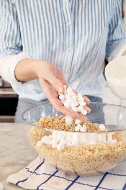 Adding mini marshmallows to rice krispies treats in large bowl.