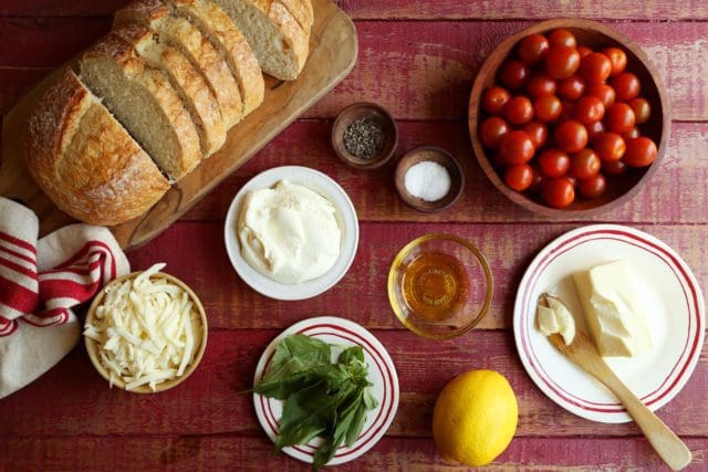 loaf of bread next to small bowls of mozzarella, ricotta, salt, pepper, oil, basil, tomatoes and butter.