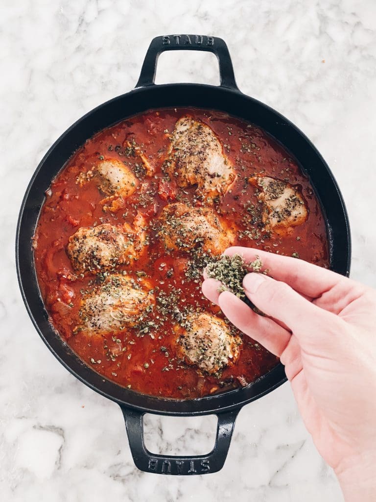 Sprinkling oregano over an easy chicken dinner recipe of tomatoes and chicken in a cast iron skillet.