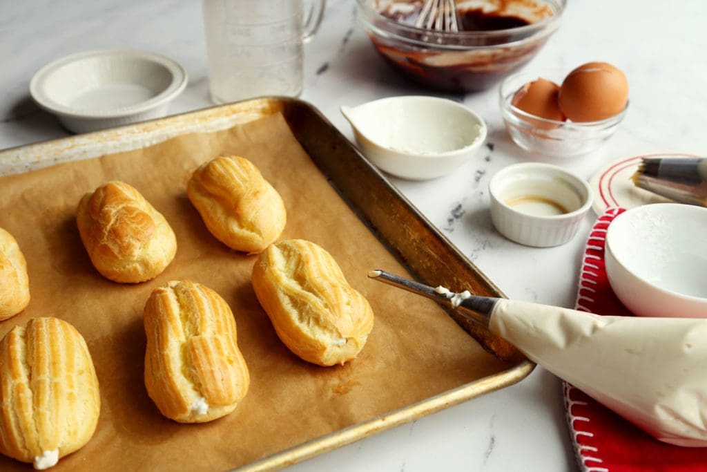 Baked eclair recipe on a baking sheet being filled with whipped cream.